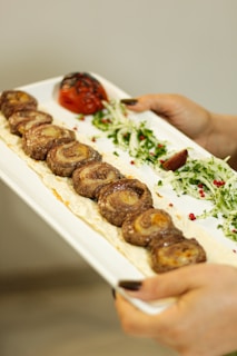 A close-up of a rectangular white plate being held, displaying a row of grilled meat rolls on a creamy sauce base. Garnished with green herbs, pink peppercorns, and sliced onions, alongside a charred tomato and a wedge of lemon. The hands with dark nail polish are visible holding the plate.
