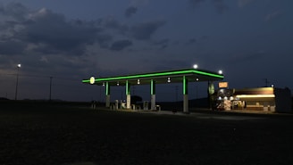 A dimly lit gas station with glowing green lights on the awning is situated against a darkening sky. To the right, a convenience store displays a bright, illuminated sign. The background features open land and scattered utility poles.
