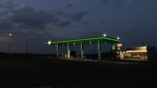 A dimly lit gas station with glowing green lights on the awning is situated against a darkening sky. To the right, a convenience store displays a bright, illuminated sign. The background features open land and scattered utility poles.