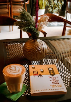 A cup of coffee with foam art rests on a green napkin next to a book titled 'Before the coffee gets cold' by Toshikazu Kawaguchi. A round vase with textured design holds various decorative grasses and plants. The scene is set on a perforated metal table in a cozy caf&eacute; environment.