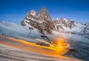 An alpine scene with snow-covered mountains under a clear evening sky. A prominent rocky peak stands tall in the center, with smaller snowy peaks in the background. In the foreground, there is a brightly lit small settlement, possibly a ski resort, casting a warm yellow light on the surrounding snow. Several ski lifts can be seen stretching up the slopes.