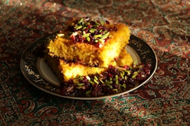 A richly decorated dish featuring golden, crispy rice topped with vibrant pistachio slivers and dark red cranberries, set on an ornate plate. The backdrop is an intricate, colorful fabric, enhancing the visual appeal.