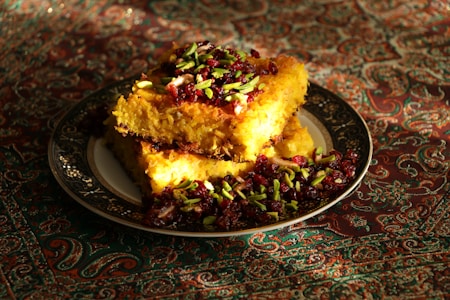 A richly decorated dish featuring golden, crispy rice topped with vibrant pistachio slivers and dark red cranberries, set on an ornate plate. The backdrop is an intricate, colorful fabric, enhancing the visual appeal.