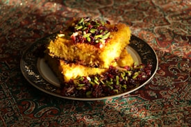A richly decorated dish featuring golden, crispy rice topped with vibrant pistachio slivers and dark red cranberries, set on an ornate plate. The backdrop is an intricate, colorful fabric, enhancing the visual appeal.
