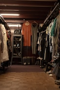 A vintage clothing store with two dresses hanging prominently in the center. The store is filled with various garments neatly hung on racks to the left and right. A glass case with jewelry is visible on the left, and a pair of old suitcases is placed on the floor. The lighting is warm, highlighting the cozy and nostalgic atmosphere.