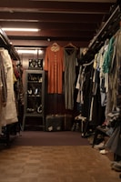 A vintage clothing store with two dresses hanging prominently in the center. The store is filled with various garments neatly hung on racks to the left and right. A glass case with jewelry is visible on the left, and a pair of old suitcases is placed on the floor. The lighting is warm, highlighting the cozy and nostalgic atmosphere.
