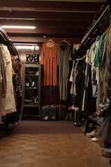 A vintage clothing store with two dresses hanging prominently in the center. The store is filled with various garments neatly hung on racks to the left and right. A glass case with jewelry is visible on the left, and a pair of old suitcases is placed on the floor. The lighting is warm, highlighting the cozy and nostalgic atmosphere.