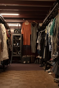 A vintage clothing store with two dresses hanging prominently in the center. The store is filled with various garments neatly hung on racks to the left and right. A glass case with jewelry is visible on the left, and a pair of old suitcases is placed on the floor. The lighting is warm, highlighting the cozy and nostalgic atmosphere.