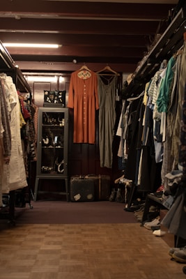 A vintage clothing store with two dresses hanging prominently in the center. The store is filled with various garments neatly hung on racks to the left and right. A glass case with jewelry is visible on the left, and a pair of old suitcases is placed on the floor. The lighting is warm, highlighting the cozy and nostalgic atmosphere.