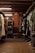 A vintage clothing store with two dresses hanging prominently in the center. The store is filled with various garments neatly hung on racks to the left and right. A glass case with jewelry is visible on the left, and a pair of old suitcases is placed on the floor. The lighting is warm, highlighting the cozy and nostalgic atmosphere.