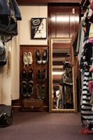 A cozy room with a wardrobe featuring hanging clothes, various shoes arranged on shelves, and a large ornate mirror resting against a burgundy door. A framed black-and-white artwork of a woman wearing a hat is displayed on the wall above the shoes. A leather bag is placed on the floor.