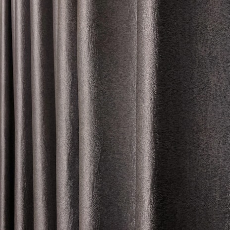 A close-up view of dark, textured drapery fabric with waves and folds creating a sense of movement. The material appears slightly shiny, indicating a smooth finish.