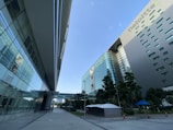 Two modern office buildings with reflective glass facades stand adjacent to each other, creating a narrow walkway between them. The architecture features sleek lines and a suspended walkway connecting the structures. Tall trees and neatly manicured landscaping are present along the pedestrian path, adding greenery to the urban scene.