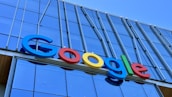 The image displays a large, colorful sign with the word 'Google' mounted on the exterior of a modern glass building. The letters are in bold, distinctly colored fonts against a backdrop of reflective windows and metallic structures.