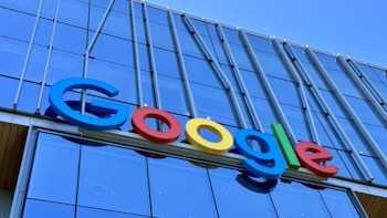 The image displays a large, colorful sign with the word 'Google' mounted on the exterior of a modern glass building. The letters are in bold, distinctly colored fonts against a backdrop of reflective windows and metallic structures.