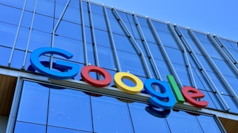 The image displays a large, colorful sign with the word 'Google' mounted on the exterior of a modern glass building. The letters are in bold, distinctly colored fonts against a backdrop of reflective windows and metallic structures.