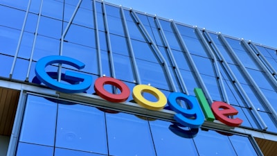 The image displays a large, colorful sign with the word 'Google' mounted on the exterior of a modern glass building. The letters are in bold, distinctly colored fonts against a backdrop of reflective windows and metallic structures.