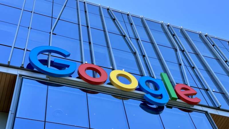 The image displays a large, colorful sign with the word 'Google' mounted on the exterior of a modern glass building. The letters are in bold, distinctly colored fonts against a backdrop of reflective windows and metallic structures.