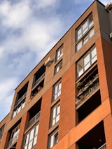 A modern apartment building with multiple stories featuring large windows and balconies adorned with plants and flowers. The building has a combination of brick and stucco exterior with a warm, earthy color palette. The image is angled upwards towards a partly cloudy sky, creating a dynamic composition.