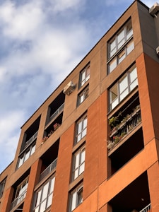 A modern apartment building with multiple stories featuring large windows and balconies adorned with plants and flowers. The building has a combination of brick and stucco exterior with a warm, earthy color palette. The image is angled upwards towards a partly cloudy sky, creating a dynamic composition.