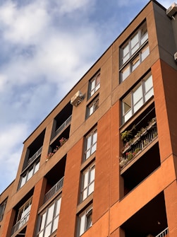A modern apartment building with multiple stories featuring large windows and balconies adorned with plants and flowers. The building has a combination of brick and stucco exterior with a warm, earthy color palette. The image is angled upwards towards a partly cloudy sky, creating a dynamic composition.