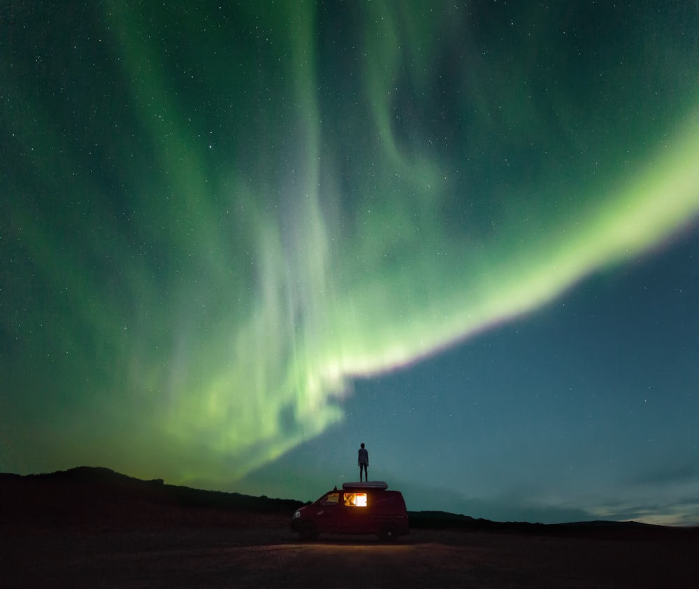 Una persona parada encima de un automóvil rojo bajo un cielo verde y azul