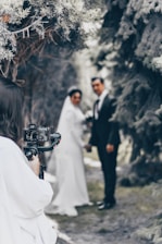 A photographer is capturing a bride and groom standing together outdoors. The couple is slightly out of focus, while the camera and photographer are closer to the viewer. Tall trees create a natural archway in the background.