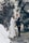 A photographer is capturing a bride and groom standing together outdoors. The couple is slightly out of focus, while the camera and photographer are closer to the viewer. Tall trees create a natural archway in the background.