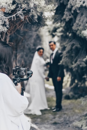 A photographer is capturing a bride and groom standing together outdoors. The couple is slightly out of focus, while the camera and photographer are closer to the viewer. Tall trees create a natural archway in the background.