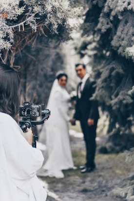 A photographer is capturing a bride and groom standing together outdoors. The couple is slightly out of focus, while the camera and photographer are closer to the viewer. Tall trees create a natural archway in the background.