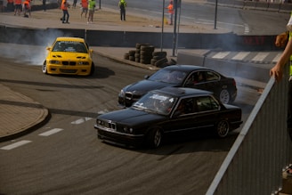 A high-speed drift race with three cars maneuvering around a corner on a racetrack. The scene is filled with smoke from the tires as the cars skillfully drift. Several people wearing orange safety vests are seen in the background, likely overseeing the event.