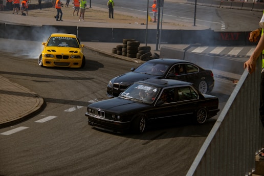A high-speed drift race with three cars maneuvering around a corner on a racetrack. The scene is filled with smoke from the tires as the cars skillfully drift. Several people wearing orange safety vests are seen in the background, likely overseeing the event.