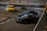 A high-speed drift race with three cars maneuvering around a corner on a racetrack. The scene is filled with smoke from the tires as the cars skillfully drift. Several people wearing orange safety vests are seen in the background, likely overseeing the event.