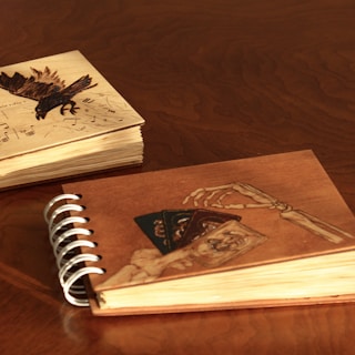 Two sketchbooks are resting on a wooden surface. The sketchbook on the left features a cover with an artistic engraving of a black bird overlying a music sheet, while the sketchbook on the right is spiral-bound and depicts a skeletal hand holding three cards with intricate designs.