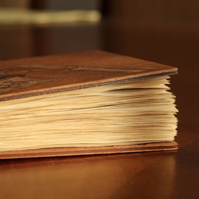 A close-up of a vintage book with a brown hard cover and slightly yellowed pages, resting on a wooden surface. The book appears to be old, with visible texture and wear on the cover.
