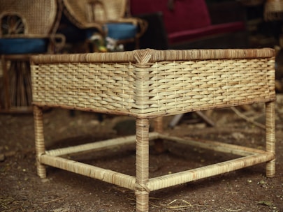 A woven rattan table stands in a rustic setting, surrounded by other wicker furniture and decorations. The texture of the rattan is prominent, with a natural, earthy tone, and the ground beneath is made of dirt, adding to the natural ambiance.
