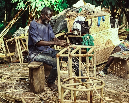 A man is sitting on a wooden stool, weaving a chair frame made of natural materials in an outdoor setting. Behind him are various wicker baskets and woven goods piled up, along with some clothing hanging over them. The environment is lush with green foliage, suggesting a rural or artisanal workspace.