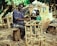 A man is sitting on a wooden stool, weaving a chair frame made of natural materials in an outdoor setting. Behind him are various wicker baskets and woven goods piled up, along with some clothing hanging over them. The environment is lush with green foliage, suggesting a rural or artisanal workspace.