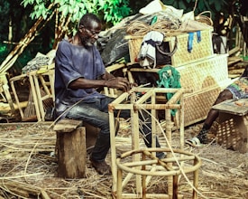 A man is sitting on a wooden stool, weaving a chair frame made of natural materials in an outdoor setting. Behind him are various wicker baskets and woven goods piled up, along with some clothing hanging over them. The environment is lush with green foliage, suggesting a rural or artisanal workspace.