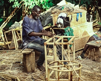 A man is sitting on a wooden stool, weaving a chair frame made of natural materials in an outdoor setting. Behind him are various wicker baskets and woven goods piled up, along with some clothing hanging over them. The environment is lush with green foliage, suggesting a rural or artisanal workspace.