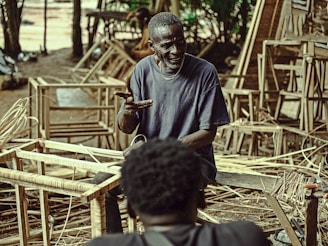 An elderly man is sitting among wooden frames and woven materials, possibly in a workshop or outdoor space. He appears to be gesturing with his hands, engaging in a conversation with another person who is partially visible in the foreground. The environment is cluttered with work materials and has a rustic, earthy feel.