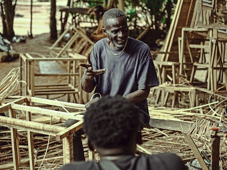 An elderly man is sitting among wooden frames and woven materials, possibly in a workshop or outdoor space. He appears to be gesturing with his hands, engaging in a conversation with another person who is partially visible in the foreground. The environment is cluttered with work materials and has a rustic, earthy feel.