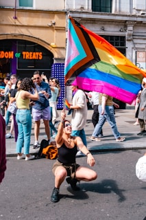 A person is joyfully kneeling in the street, holding a large rainbow flag with additional colors representing inclusivity. Behind them, a diverse crowd is gathered, some taking photos or interacting with each other. The scene is set in an urban environment with visible storefronts and people walking by.