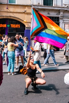A person is joyfully kneeling in the street, holding a large rainbow flag with additional colors representing inclusivity. Behind them, a diverse crowd is gathered, some taking photos or interacting with each other. The scene is set in an urban environment with visible storefronts and people walking by.