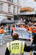 A group of people participate in a parade on a city street, holding a banner with messages promoting love, unity, and LGBTQ+ rights. They are surrounded by onlookers behind barriers, and a security guard stands in the foreground with a high-visibility jacket. Buildings and temporary structures are visible in the background.