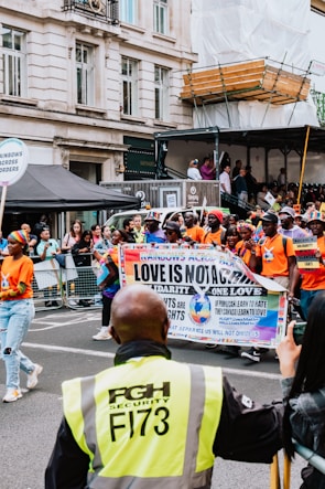 A group of people participate in a parade on a city street, holding a banner with messages promoting love, unity, and LGBTQ+ rights. They are surrounded by onlookers behind barriers, and a security guard stands in the foreground with a high-visibility jacket. Buildings and temporary structures are visible in the background.