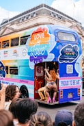 A colorful double-decker bus with signage promoting Pride and inclusivity is parked on a city street. Multicolored advertisements, including logos from various sponsors, decorate the side. A person dressed in vibrant, expressive clothing and high heels is seen interacting with the bus at its open rear entrance. A crowd is gathered around, showcasing a lively and festive atmosphere.