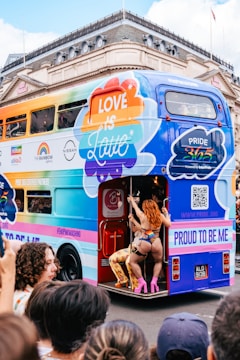 A colorful double-decker bus with signage promoting Pride and inclusivity is parked on a city street. Multicolored advertisements, including logos from various sponsors, decorate the side. A person dressed in vibrant, expressive clothing and high heels is seen interacting with the bus at its open rear entrance. A crowd is gathered around, showcasing a lively and festive atmosphere.