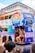 A colorful double-decker bus with signage promoting Pride and inclusivity is parked on a city street. Multicolored advertisements, including logos from various sponsors, decorate the side. A person dressed in vibrant, expressive clothing and high heels is seen interacting with the bus at its open rear entrance. A crowd is gathered around, showcasing a lively and festive atmosphere.