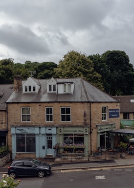 A two-story building made of stone bricks, featuring small businesses on the ground floor including a personal training center, a coffee shop with outdoor seating, and an interior design store. The sky is overcast with clouds, and there is a parked car on the street in front of the building. Lush green trees are visible in the background.
